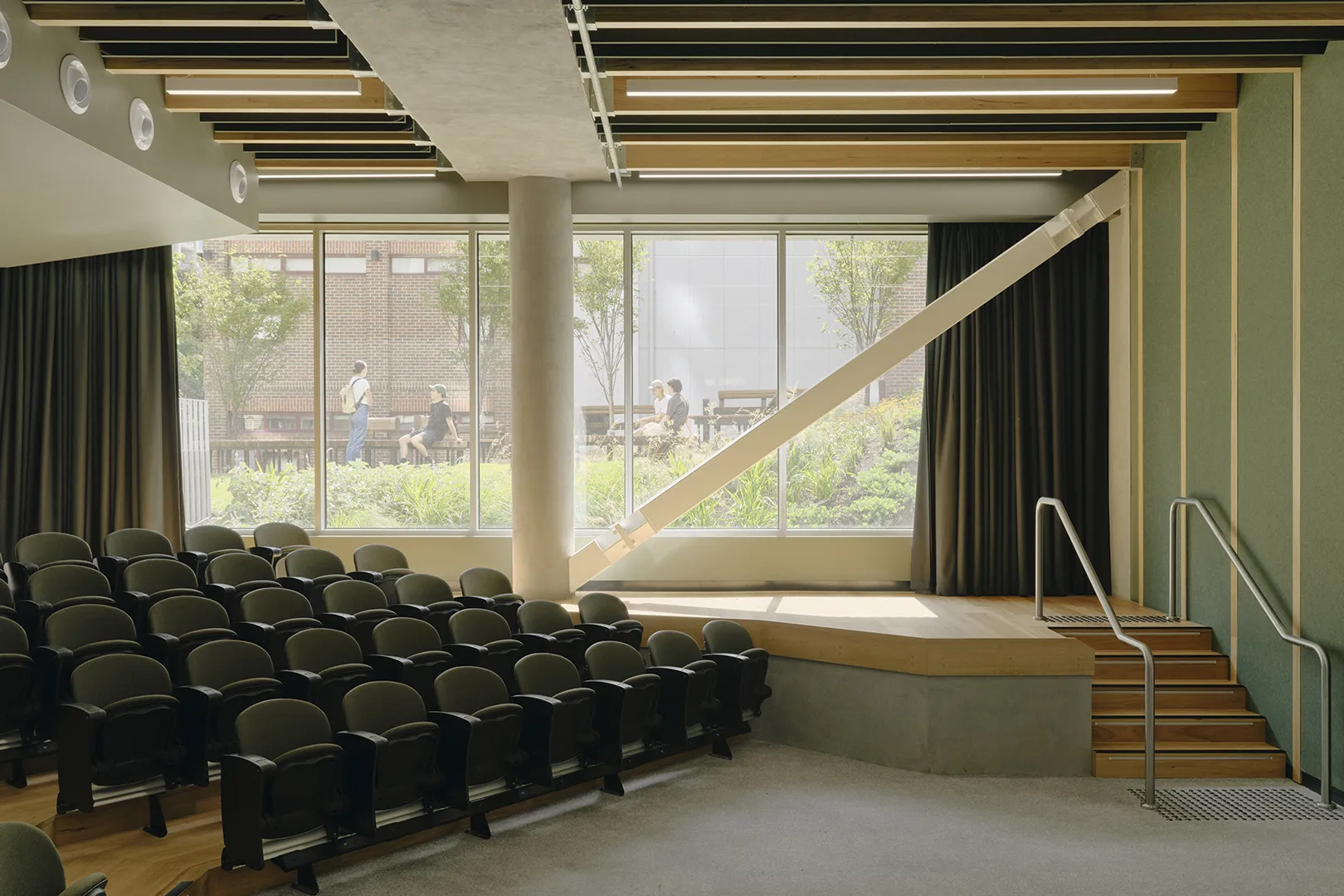 Auditorium with tiered seating, a presentation stage, and large windows overlooking outdoor gardens.