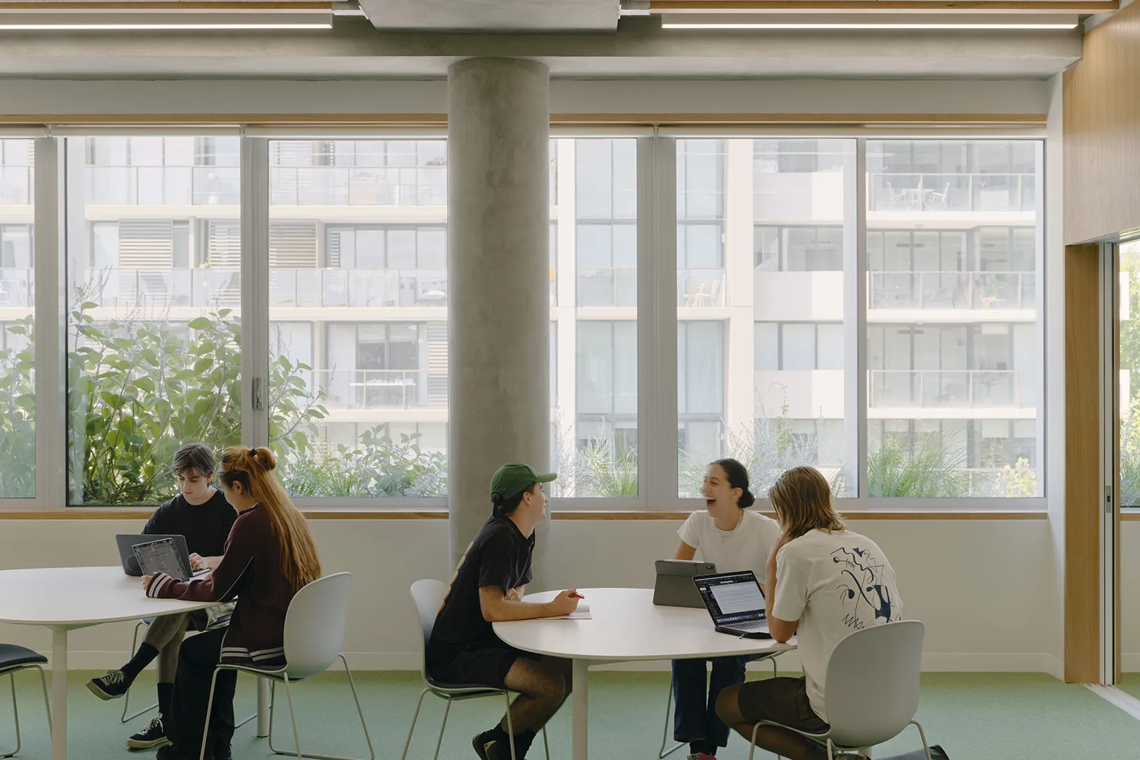 Students collaborating at tables in a shared learning space with large windows and natural light.