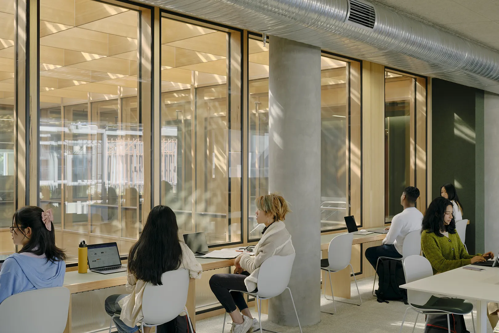 Students studying at individual workstations beside large windows with views across the building’s shared spaces.