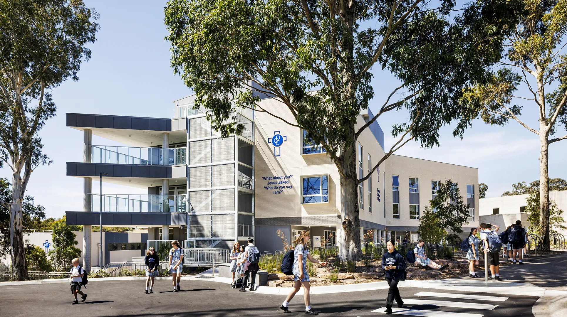 Students gathering outside the school building with landscaped gardens, outdoor walkways, and shaded campus spaces.