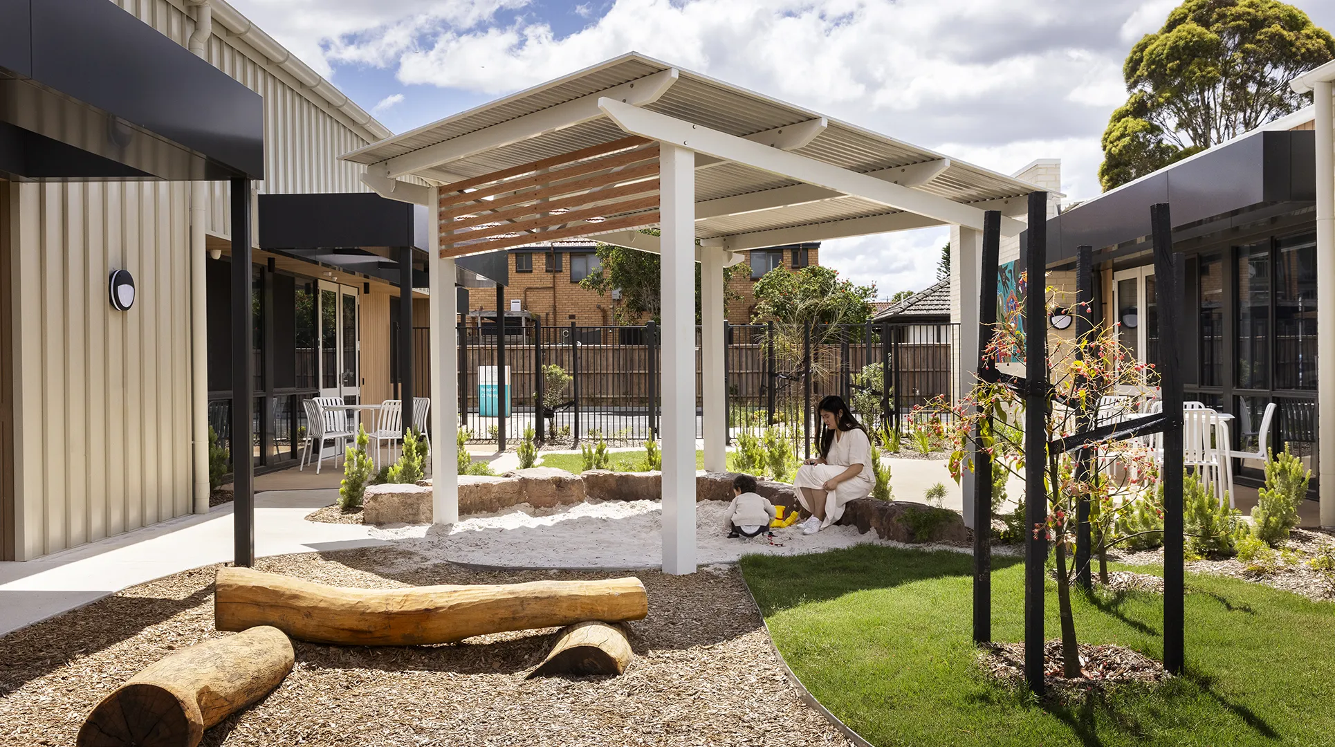 Outdoor courtyard with shaded play areas, landscaped gardens, and space for families and young children to gather and explore.