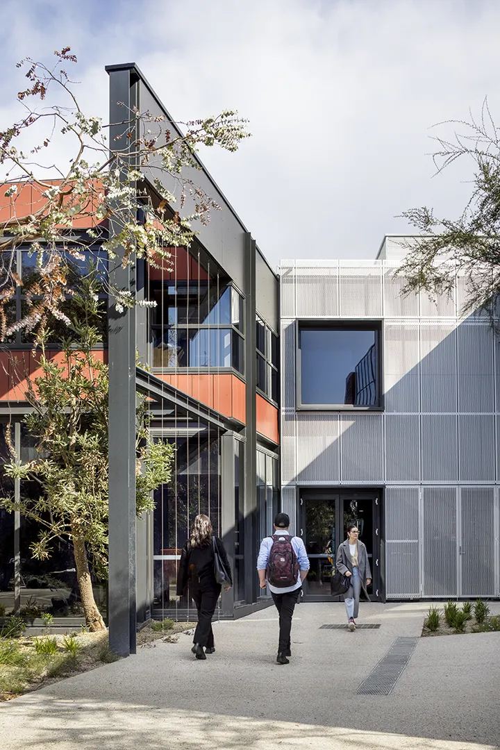 People entering and leaving a campus building with large windows, outdoor pathways, and landscaped planting.