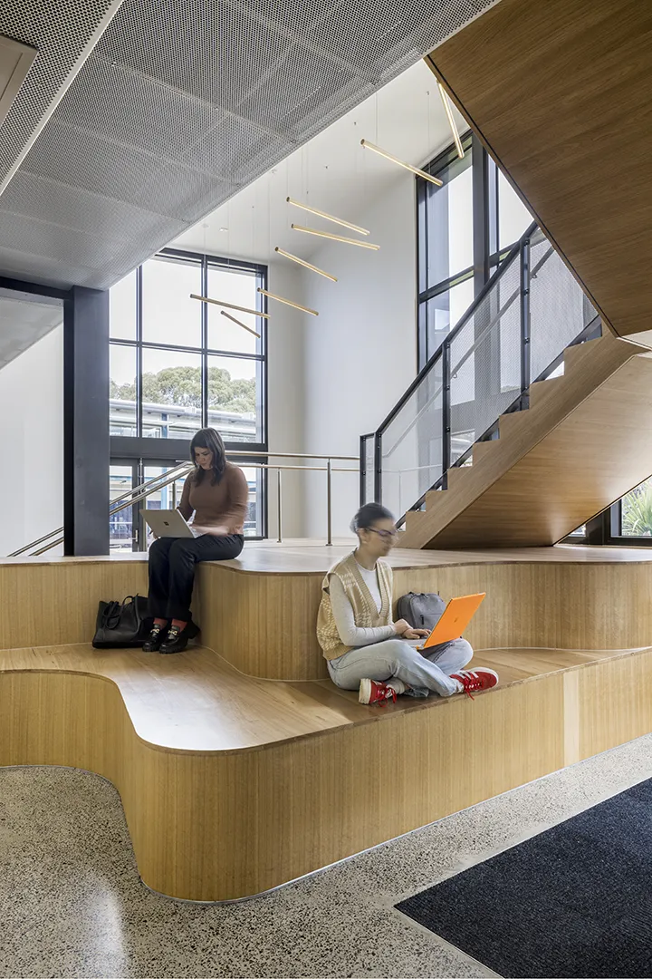 People using tiered seating beneath a staircase in a shared study and collaboration space with large windows and natural light.