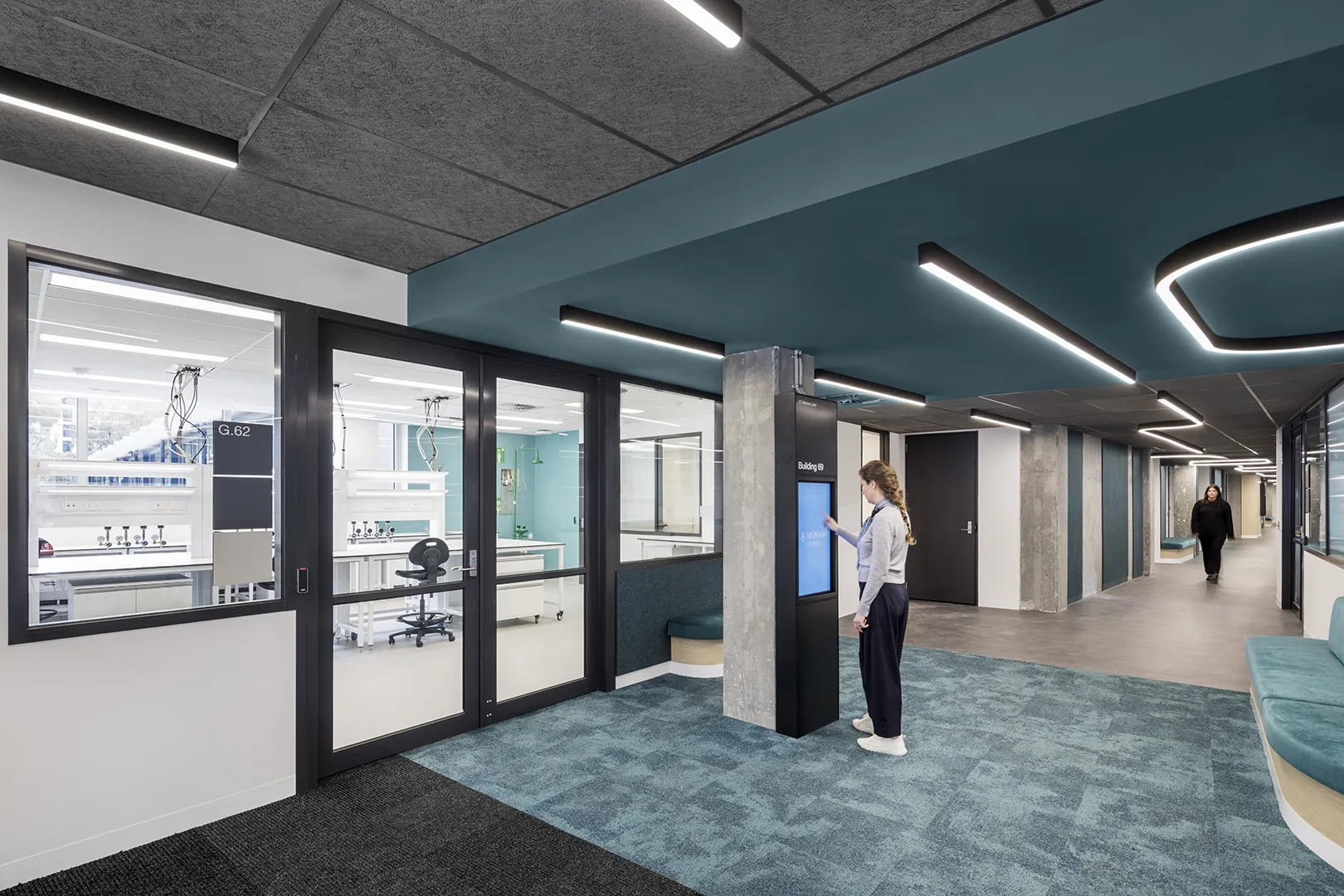 Building corridor with glass-fronted laboratories, informal seating, and digital wayfinding for learning and research spaces.