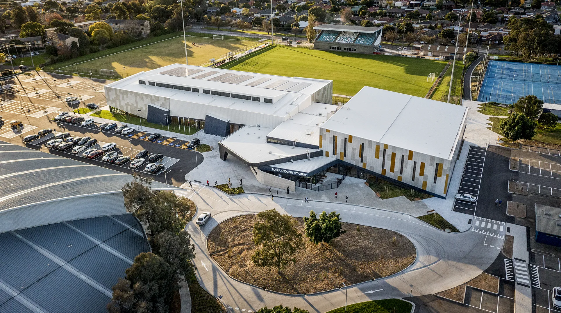 Aerial view of the stadium and surrounding sports precinct, including playing fields, tennis courts, parking areas, and pedestrian access.