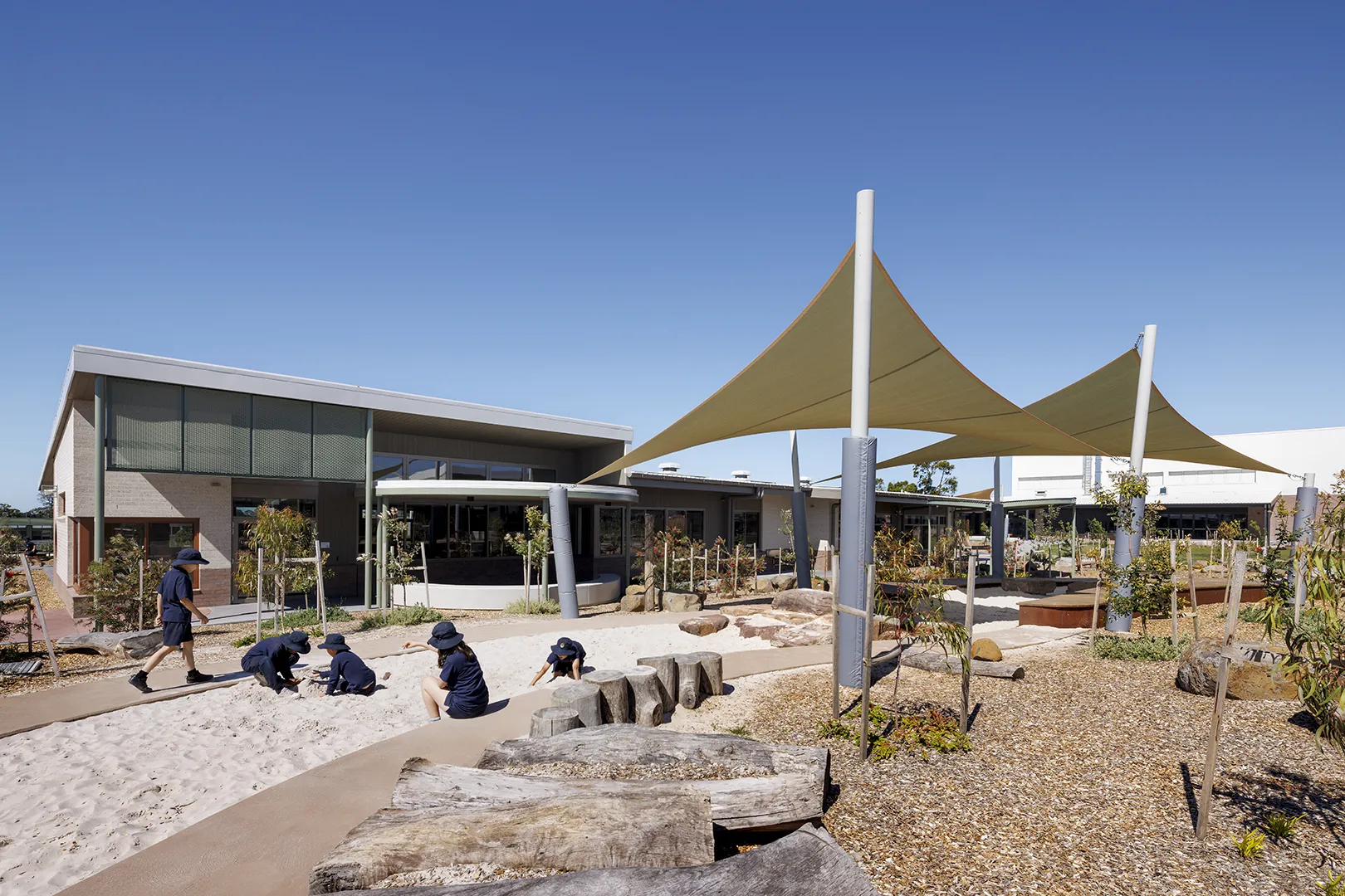 Outdoor learning and play area with tensile shade structures, landscaped gardens, natural play elements, and adjacent classroom buildings.