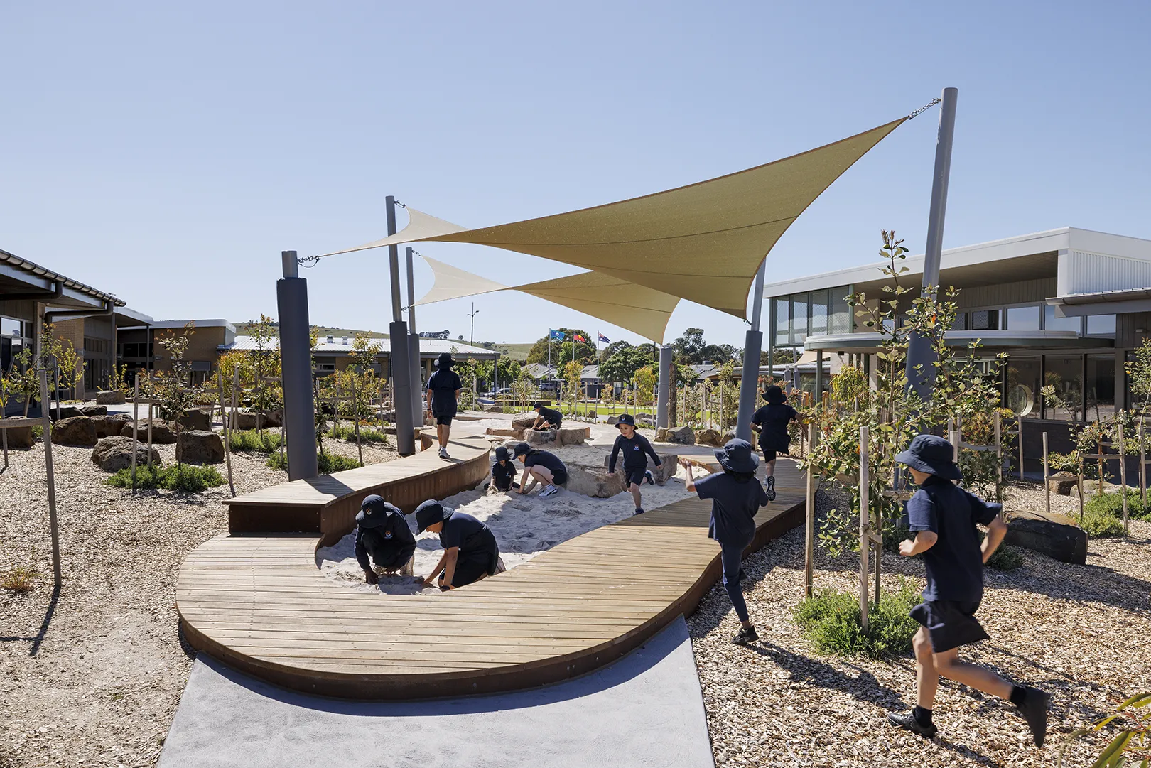 Children using a shaded outdoor play space with sand, timber seating, pathways, and landscaped gardens.