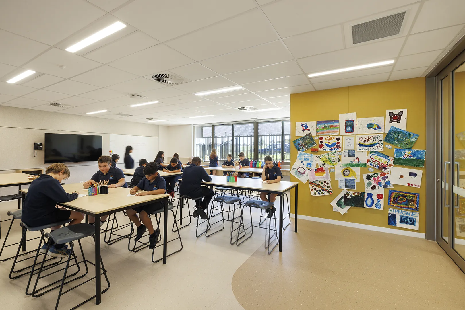 Students working at tables in a classroom with displayed artwork, large windows, and flexible learning spaces.