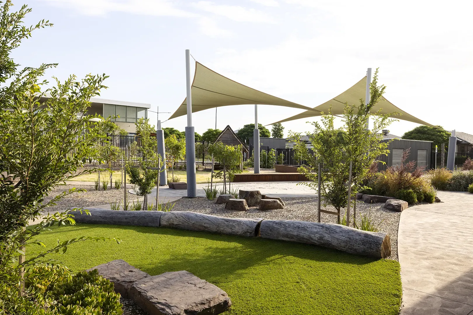 Landscaped outdoor play area with shade sails, natural play elements, seating, and winding pathways.