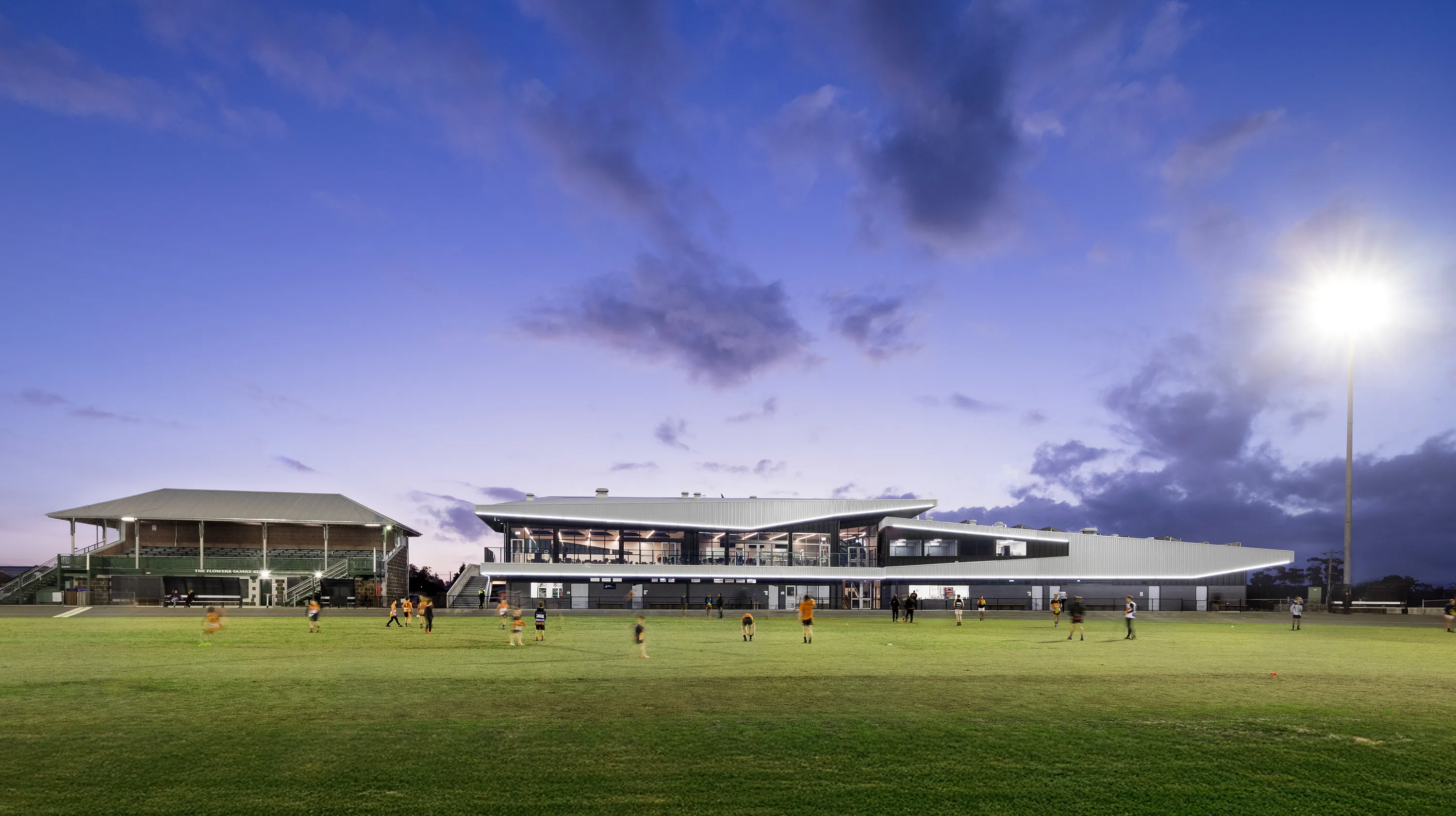 Sports ground at dusk with community members using the oval and illuminated facilities in the background.